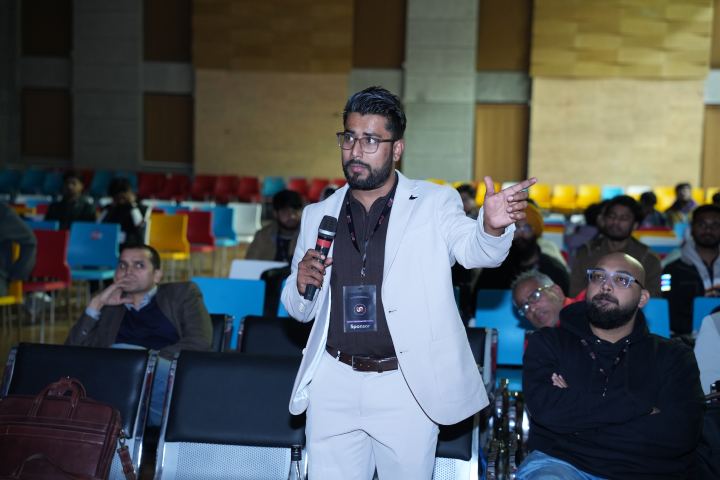 A standing attendee in a cream blazer holding a microphone in one hand and gesturing with the other while making a point, rows of red, blue and yellow seats and a dozen or so seated listeners behind them.