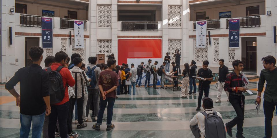 Around forty people queueing in a loose line across a tall atrium towards a registration table at the far wall, most of them carrying backpacks, tall printed banners of speaker portraits standing down both sides and a lit screen on the wall behind the desk.