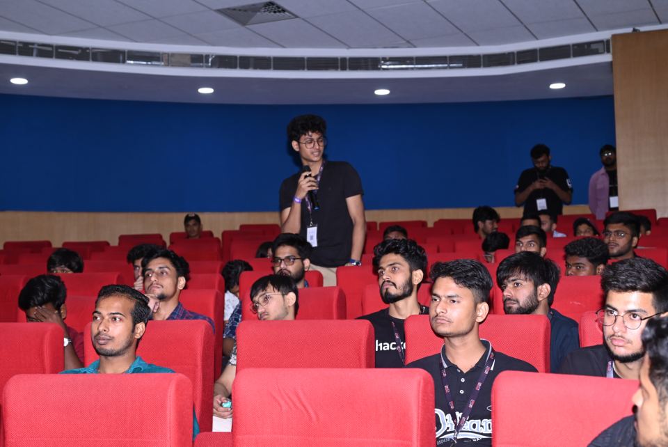 A raked auditorium of red seats seen from the side, one person standing in a middle row with a handheld microphone part-way through a question, around fifteen more seated in front of them all facing a stage outside the frame, several of them wearing lanyards, and two more standing against the back wall.