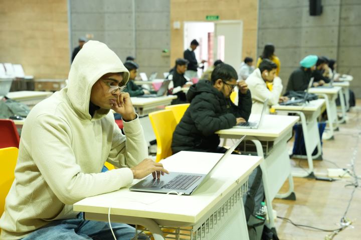 A player in a cream hooded top leaning in close to a laptop with one hand at their glasses, and a line of desks receding behind them where six or seven more people work at laptops under concrete pillars.