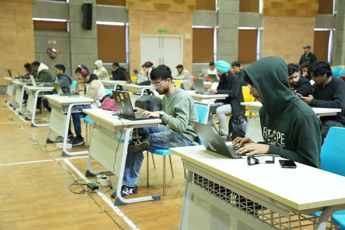 A wide view down a long line of white bench desks in a hall with a pale wooden floor, around twenty-five people working at laptops with headphones and phones beside them, teal and yellow chairs, taped cable runs across the floor and a green fire exit sign above double doors at the far end.