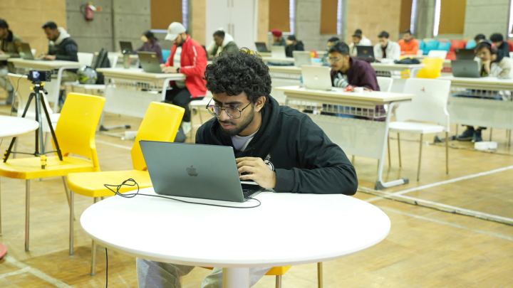 One laptop from over the player's shoulder, a dark terminal filling the screen, with a second competitor at the next desk along and the hall out of focus behind.