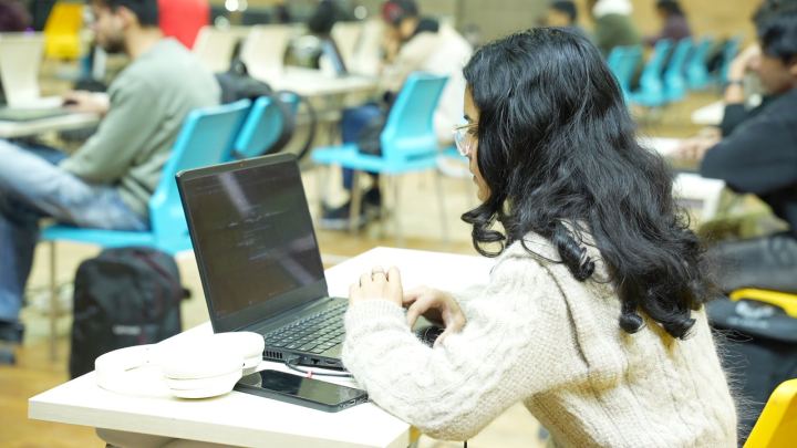 A side table against a window wall, two people working on laptops with a third leaning over to look, daylight coming in behind them.