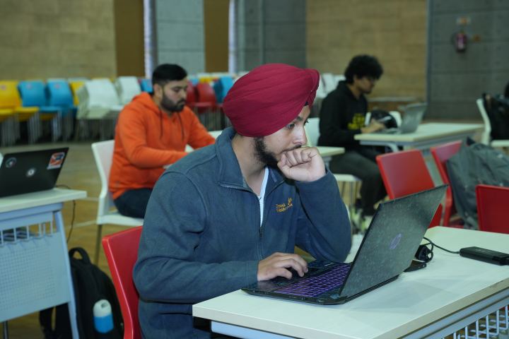 Two players sharing one long desk, the nearer one in a navy fleece with a laptop open on a terminal, the further one watching their screen, and a paper cup and a phone between them.