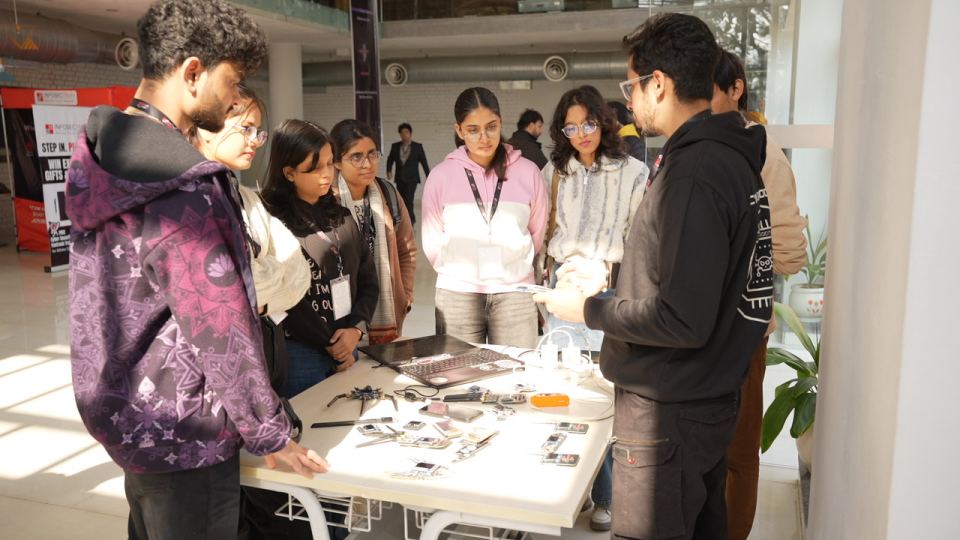 A ring of people standing shoulder to shoulder around a white table in a bright hall, all of them looking down at it, the table covered with opened-up handsets, a laptop and small hand tools, with a printed roll-up at the left edge of the frame.