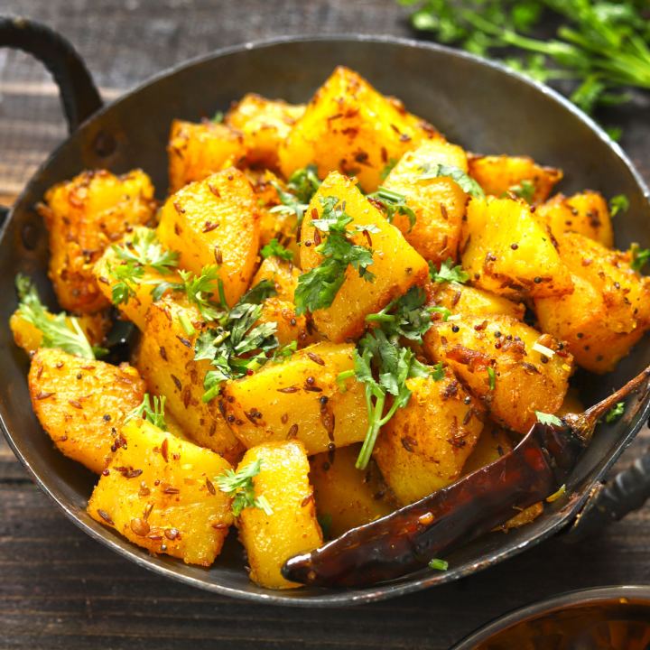Cubes of potato fried yellow with cumin and mustard seed, scattered with coriander and laid with a whole dried red chilli, in a dark iron pan on a wooden table.