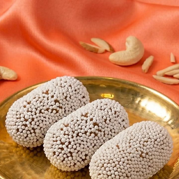 Three pieces of bal mithai, brown fudge rolled in tiny white sugar balls, in a hammered brass dish on orange cloth with cashews and slivered almonds scattered behind.