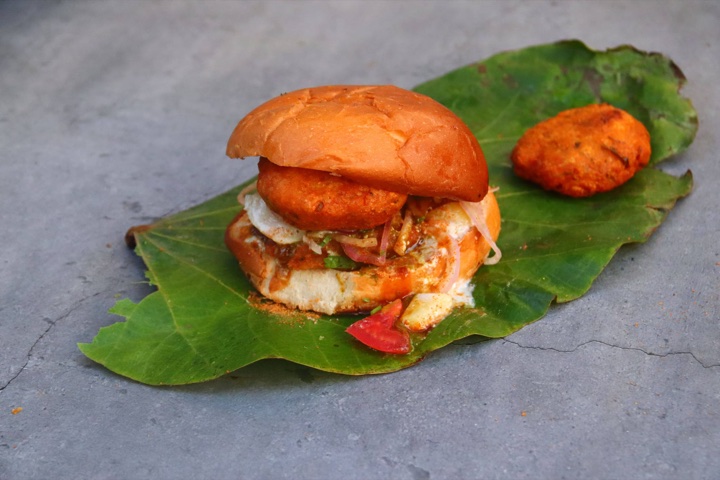 A bun tikki filled with a fried potato patty, chutney, raw onion and tomato, served on a green leaf with a second tikki behind it.