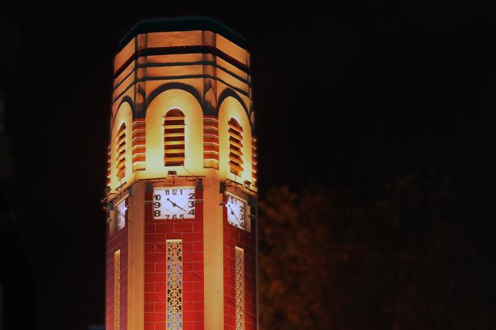 The upper stage of a many sided brick clock tower floodlit gold against a black sky, arched louvred openings above a band of white clock faces, with unlit foliage on the right.