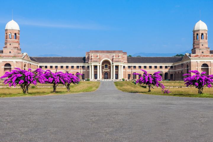 A long symmetrical brick building with a central arched portico and a domed tower at each end, seen straight down a wide driveway lined with bougainvillea in full magenta flower under a clear blue sky.