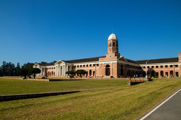 A long colonial building in pink brick and cream, its white domed tower rising over a columned portico and a run of arches, seen across a wide lawn with a few people sitting along its edge.