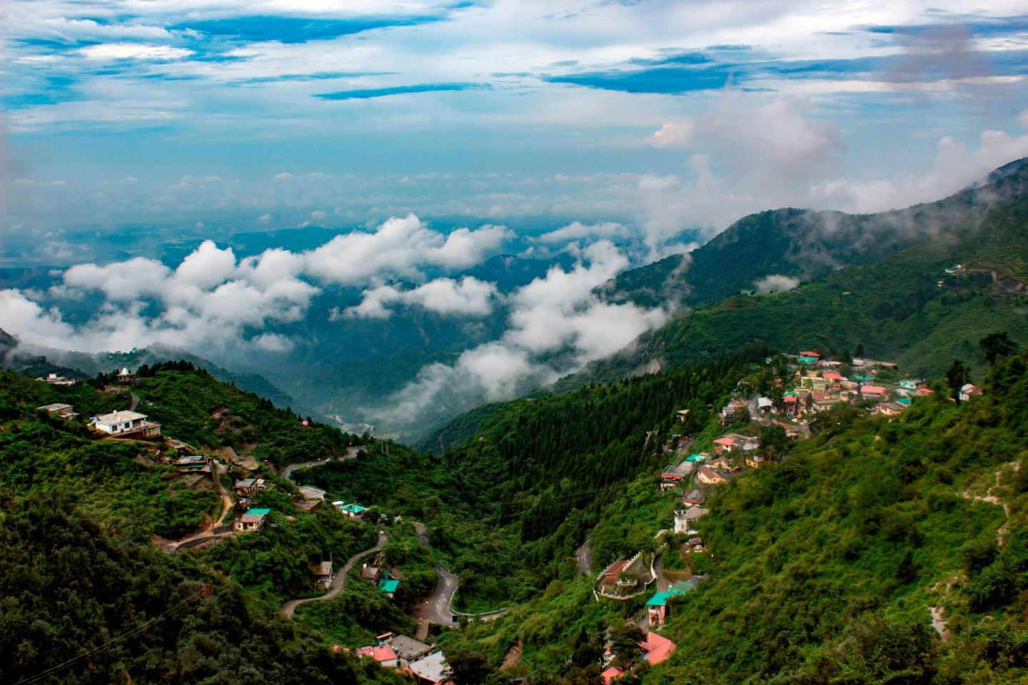 The Doon valley seen from a forested ridge above it, a hairpin road climbing away on the left, roofs stacked down the slope on the right, low cloud lying across the middle distance and the plain beyond it.