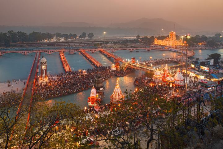 The bathing ghats at Haridwar from above at dusk, the stepped banks and footbridges dense with people, a clock tower on the near bank and lit temple spires along the water.