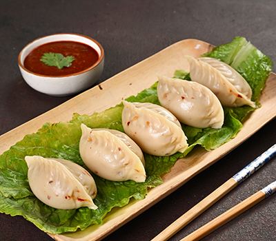 Five pleated steamed momos in a row on a lettuce leaf on a pale wooden platter, a small white bowl of dark red chutney with a coriander leaf on it behind them, and a pair of blue patterned chopsticks lying on the dark table alongside.