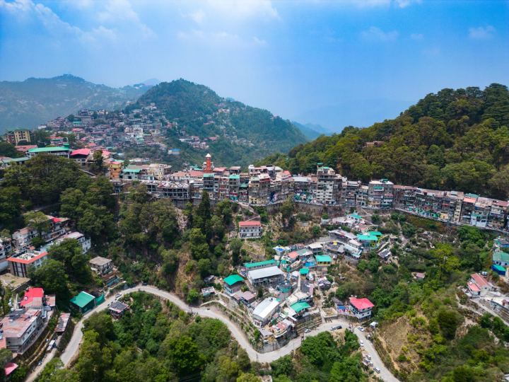 Mussoorie seen from the air, its buildings stacked in tiers along the top of a ridge above a long retaining wall, tin roofs in green and red, a square orange tower with a pale pyramid roof standing above them, a road switchbacking down through the trees below the town, and further ridges going back into haze.