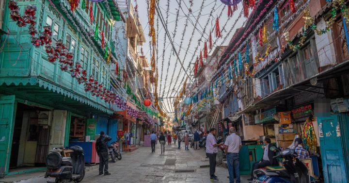 A narrow market street strung across with tinsel and paper streamers, green wooden balconies over the shopfronts on the left, stalls and boards on both sides, scooters parked along the edge and people walking down the middle.