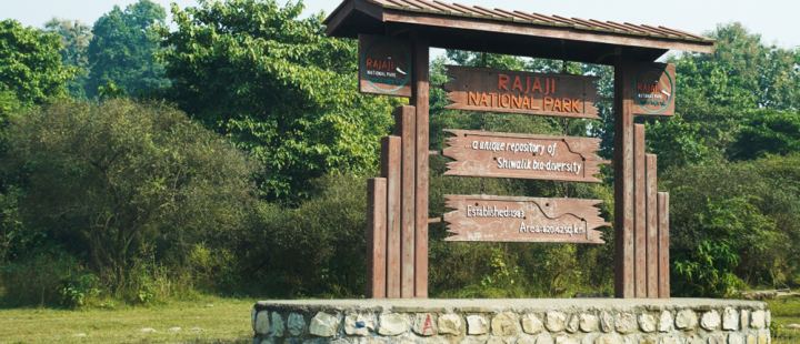 A wooden park gate across a road, its name-board on the crossbeam and two painted signs hanging under it, with scrub and tall grass closing in behind.