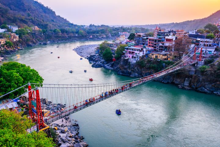 A red steel suspension footbridge crossing a wide green river, people walking across it, rafts on the water below and a town stacked up the hillside on the far bank.