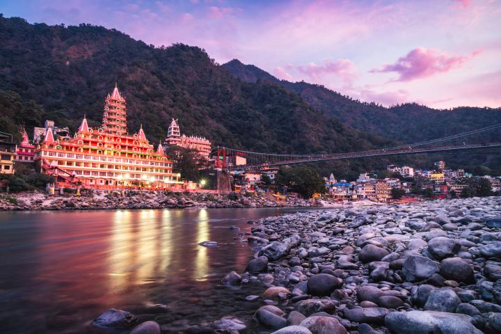 A wide river at dusk with a many tiered temple lit gold on the far bank, a suspension footbridge crossing to a town stacked up the hillside, forested hills behind and pale boulders in the foreground under a pink and violet sky.