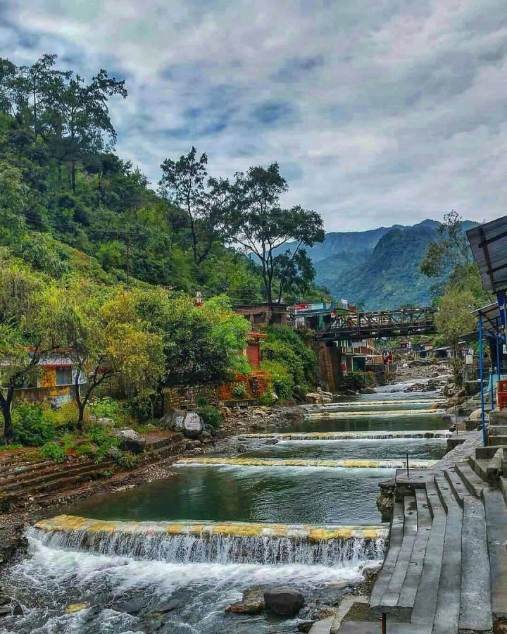 A chain of low stone weirs stepping a stream down a valley floor, each spilling into a still green pool, with cut stone bathing steps along the near bank and forested hills behind.