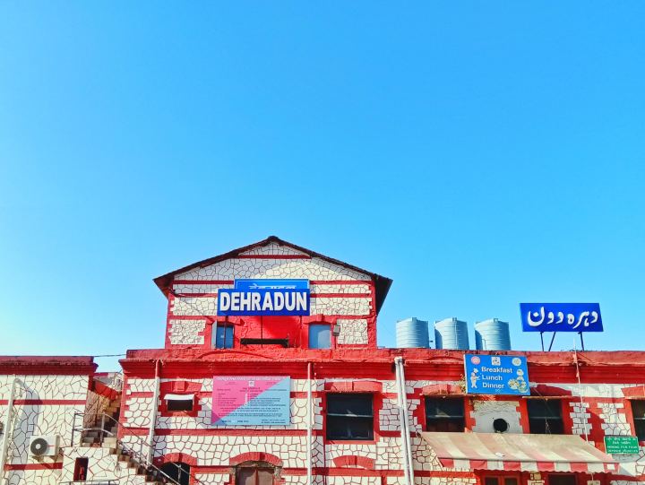 The terminus building at Dehradun station, faced in red and white crazy paving, its name on blue boards in Devanagari, Latin and Urdu script, with water tanks along the roofline under a clear sky.