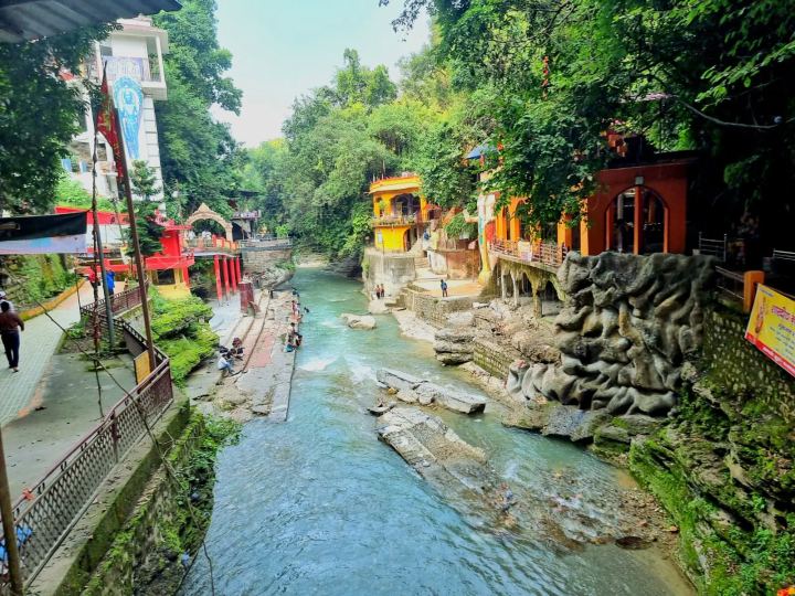 A shallow river running between built-up banks in a wooded gorge, an ochre and red temple complex set into the rock on the right, a painted gateway and walkway on the left, and people sitting on the stone bank.