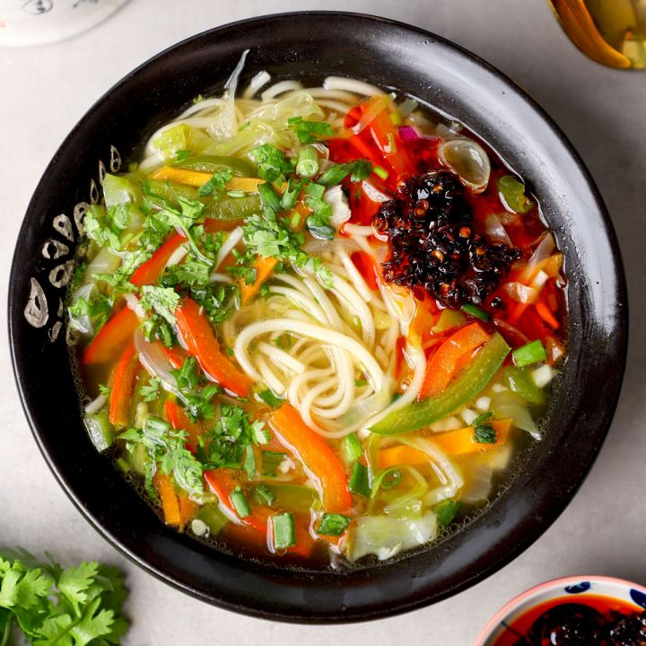 A dark bowl of thukpa shot from above, wheat noodles coiled in a clear broth with strips of carrot and green pepper, shredded cabbage, coriander and spring onion, and a spoonful of chilli oil on top.