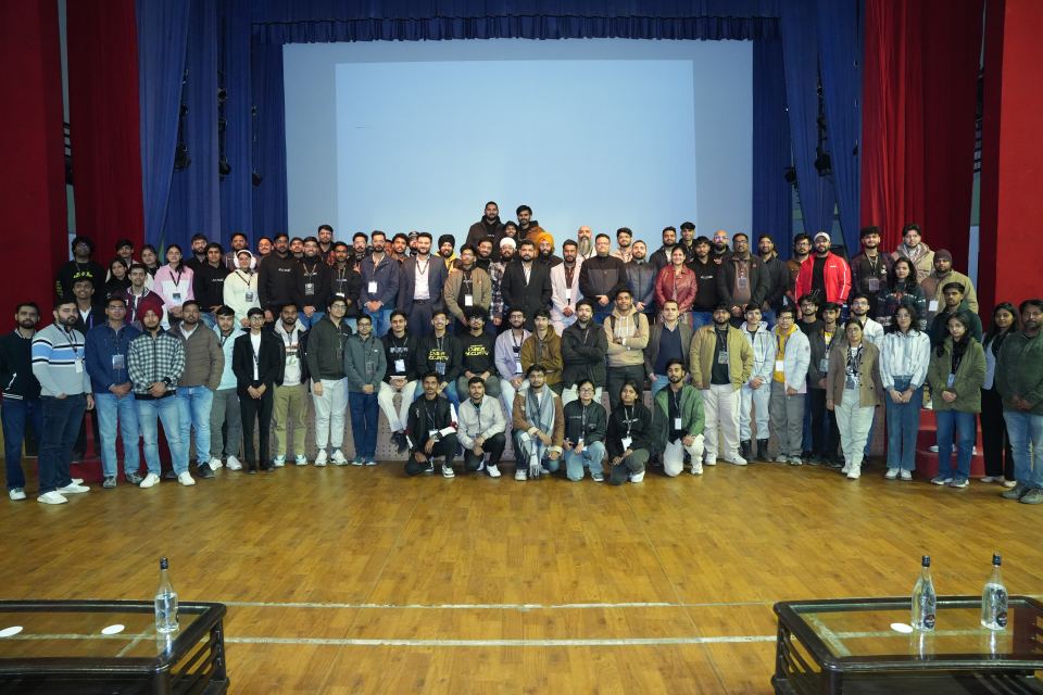 Close to a hundred people packed into four deep rows across a wide wooden stage for a group photograph, most of them wearing lanyards, a blank lit projection screen above them and heavy red and blue stage curtains down both sides.