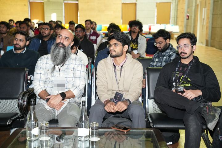 Three attendees in the front row on black leather chairs, listening, a low glass table of water bottles and glasses in front of them and thirty or so more people seated in the rows behind.