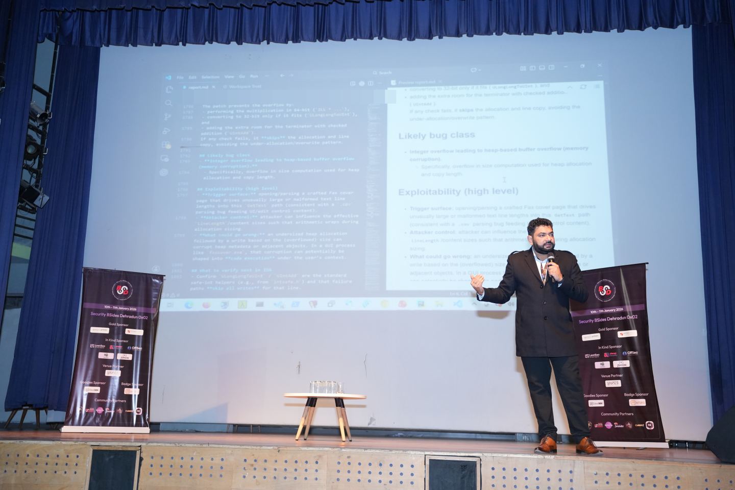 A wide hall with a speaker standing at the front beside a large projected document, a full-height printed sponsor roll-up standing at each side of the stage with every tier of names legible on both, an empty stage front in the foreground and rows of seats beyond.