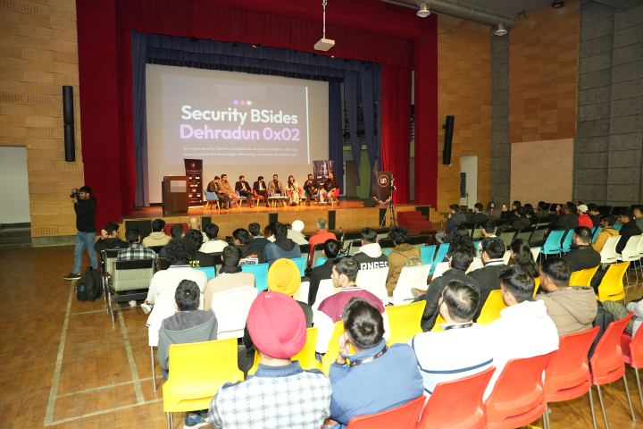 A hall seen from the back row, sixty or seventy people seated in yellow, orange and teal chairs facing a lit stage where eight panellists sit in front of a large projected title slide, a wooden lectern at one side of the stage and a photographer standing in the left aisle.
