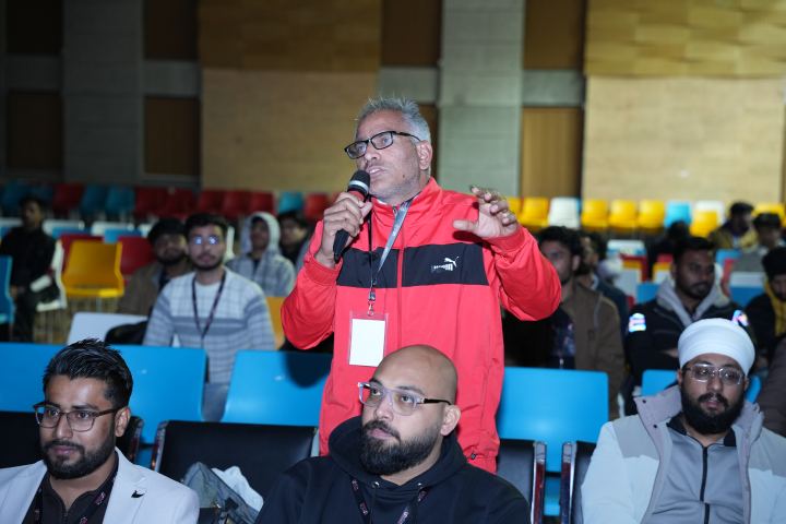 A standing attendee in a red jacket speaking into a microphone with their other hand raised open as they ask a question, three people seated in the row in front of them and around fifteen more behind.