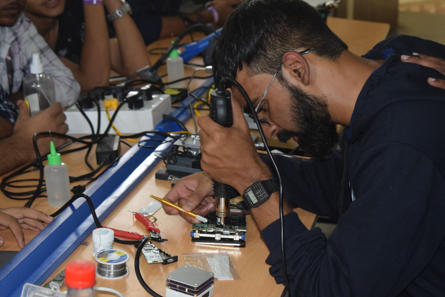 Someone bent low over a workbench with a hot-air rework tool held at their eye and a fine probe in the other hand, working on a small green circuit board clamped in a jig. Flux bottles, a reel of solder, cutters and loose boards are spread along the bench, and other people lean in from the left.