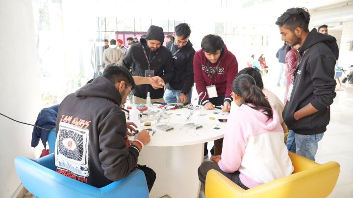 Eight or nine people crowded round a white circular table in a glass-walled atrium, most of them standing and leaning in over small boards, tools and a water bottle spread across the middle, with blue and yellow tub chairs pulled up to it.