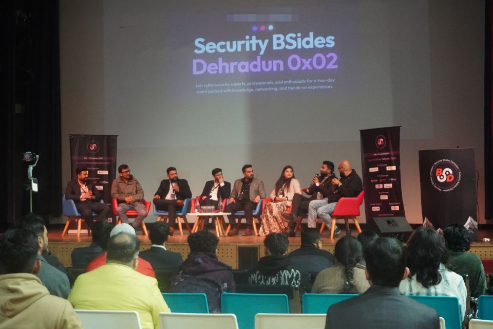 Eight people sitting in armchairs across a low stage, one of them speaking into a handheld microphone, a large projected title slide on the wall above them, a printed sponsor roll-up standing at each end of the stage, a lectern at the right carrying the conference's own roundel, and the backs of the audience's heads filling the bottom third of the frame.