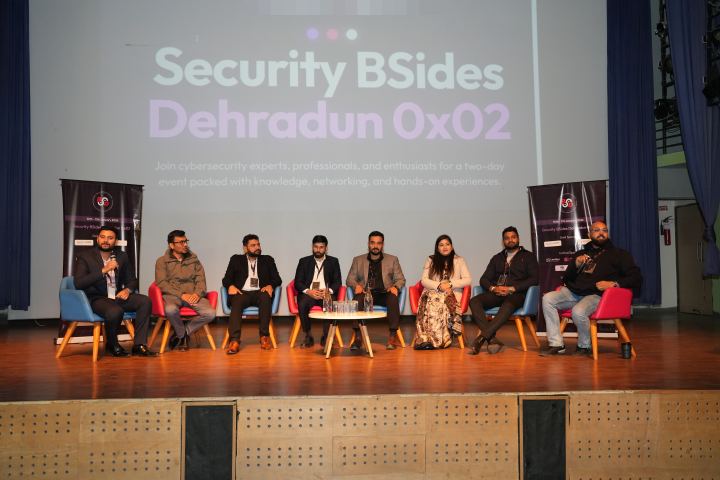 Eight panellists seated in a line of coloured armchairs on a stage around a low round table of water glasses, the one at the left end speaking into a handheld microphone, a projected title slide filling the wall behind them and a printed roll-up banner standing at each end of the stage.