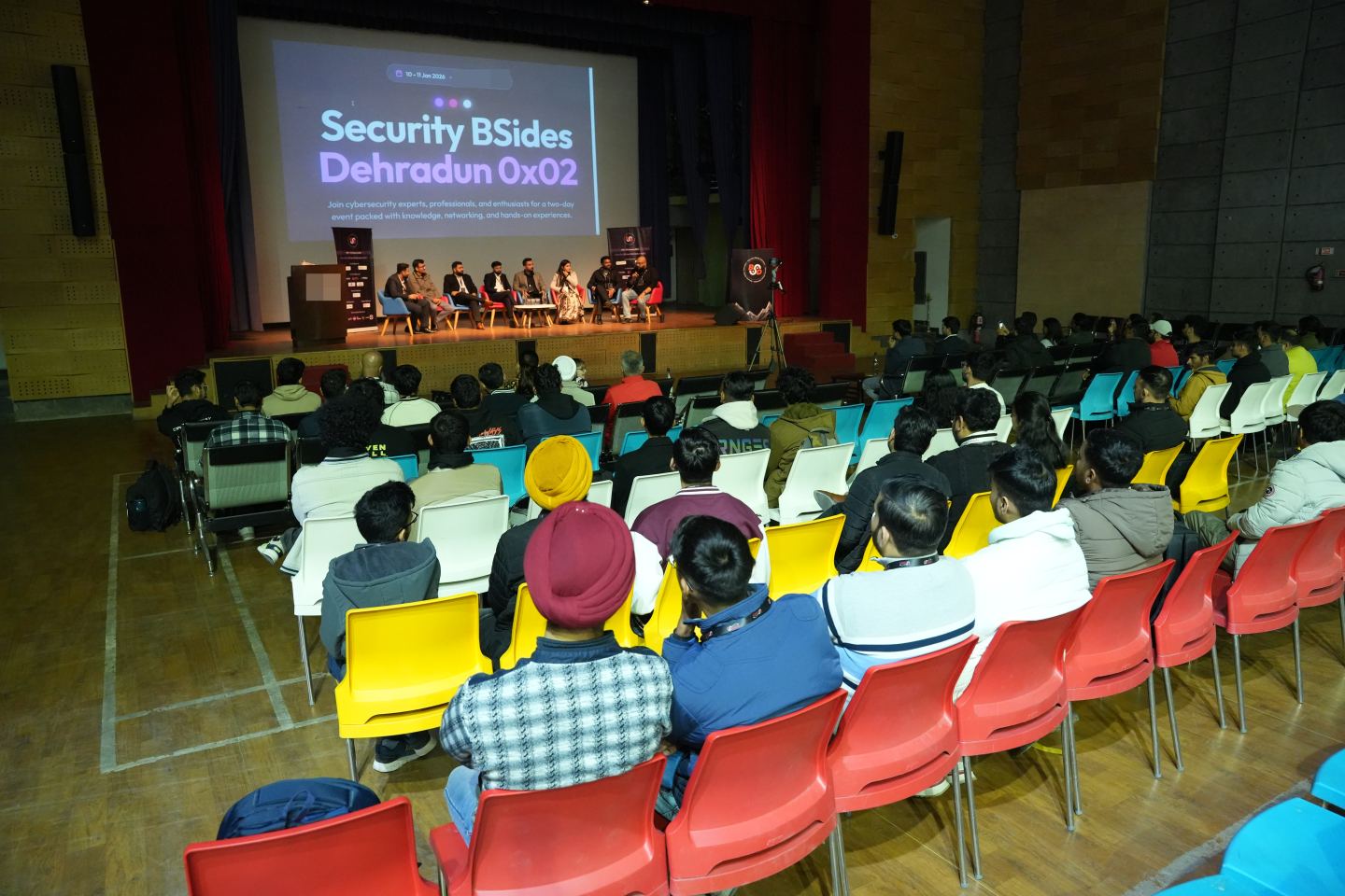 Eight panellists in a row of chairs on a lit stage, one of them speaking into a handheld microphone, a projected title slide filling the wall behind them, a printed roll-up banner at each end of the stage and the backs of the audience's heads across the bottom of the frame.