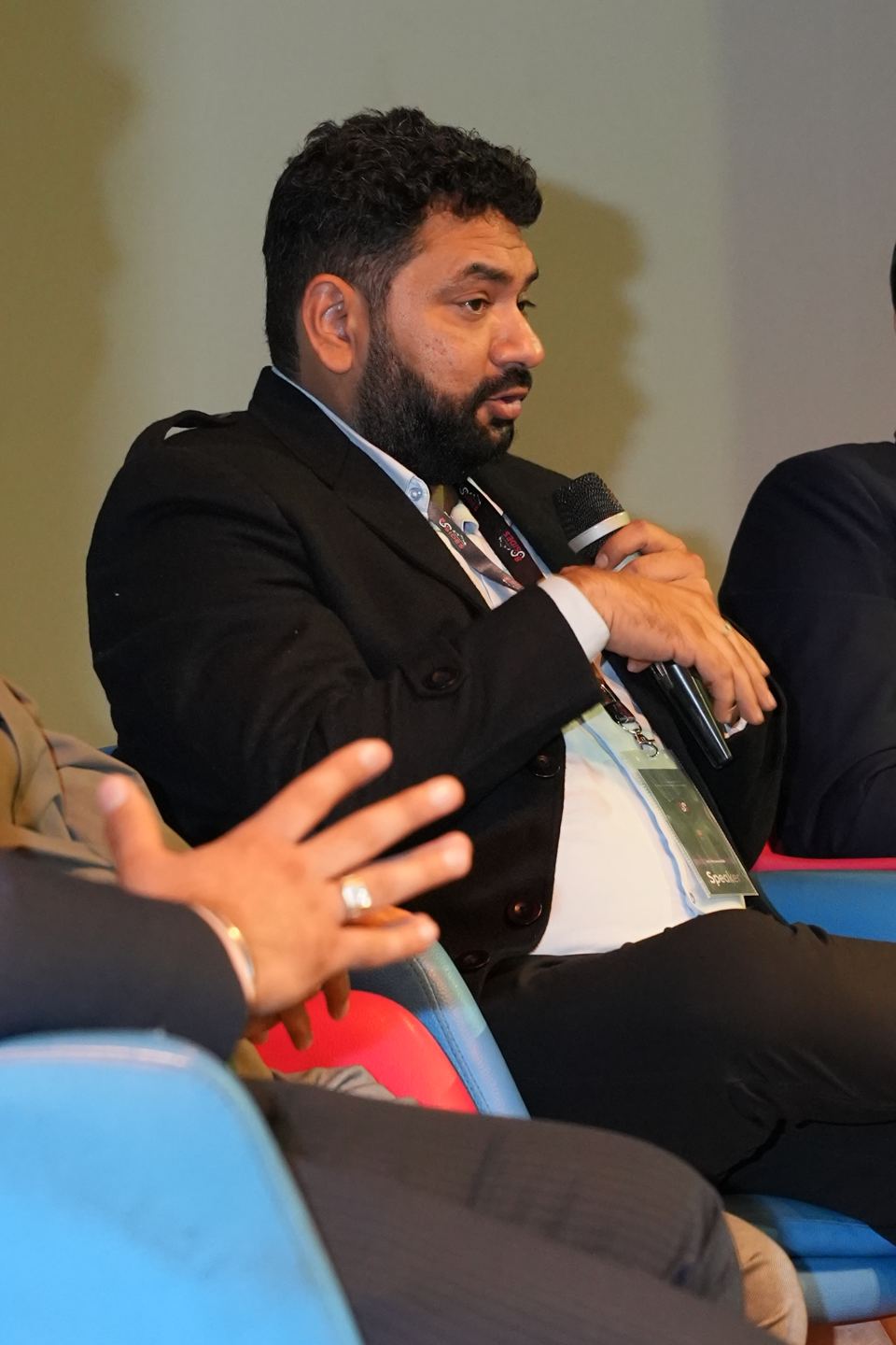 Somebody sitting forward in a low chair on a panel, part-way through an answer, holding a handheld microphone up in one hand, wearing a dark jacket over a white shirt with a lanyard and badge card round their neck, with the open hand of the person beside them in the foreground.