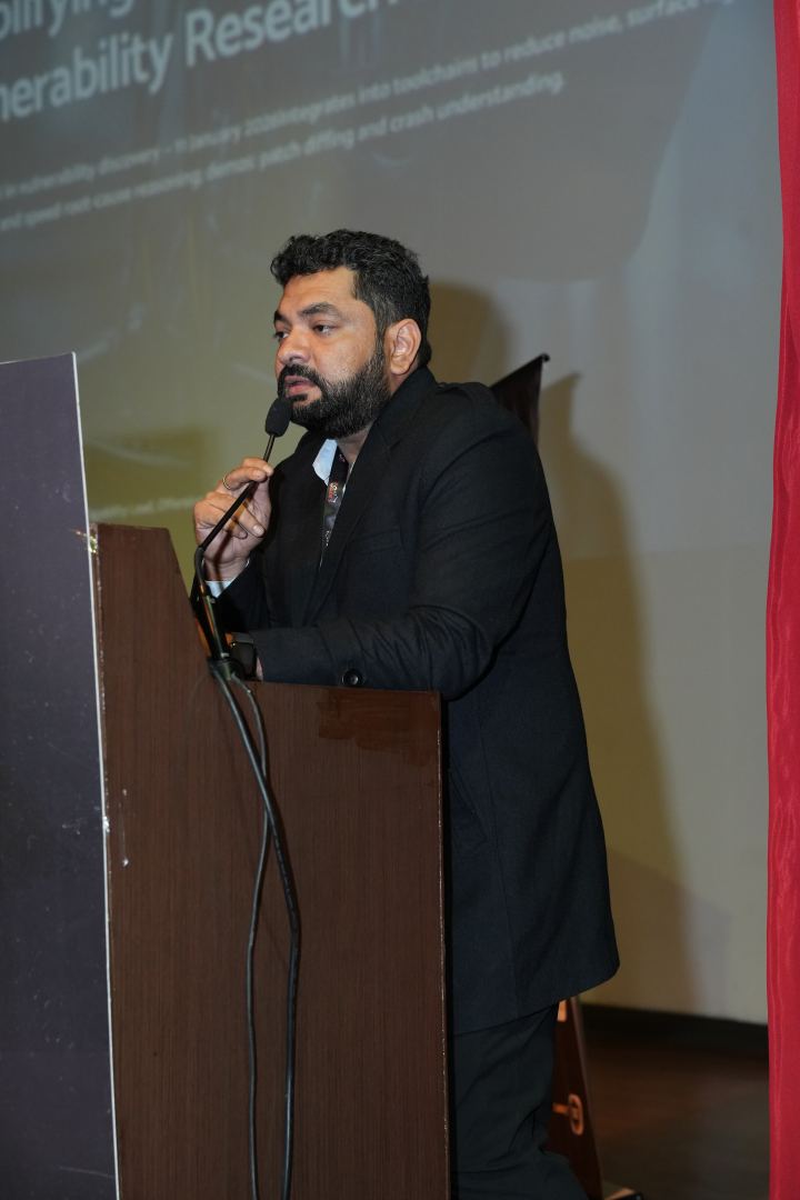 A speaker standing behind a wooden lectern with a gooseneck microphone bent up towards them, one hand raised beside it, wearing a dark jacket and a lanyard, with a projected title slide on the wall behind and a heavy red curtain at the edge of the frame.