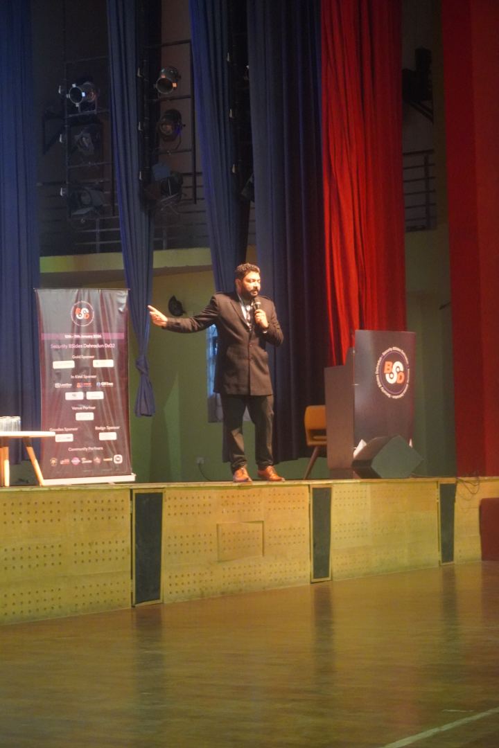 A speaker alone on a wide stage in a suit jacket, handheld microphone in one hand and the other arm open in a gesture, blue and red stage curtains and a lighting truss behind them, a full printed sponsor roll-up standing at the left and a lectern at the right.