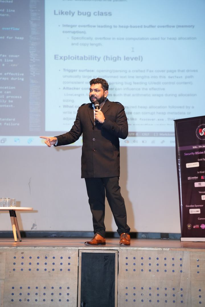 A speaker standing at the front of a stage pointing off to one side, microphone in the other hand, a projected document of headed notes and bullet points filling the wall behind them, a small table of water glasses at the left and a printed sponsor roll-up at the right.