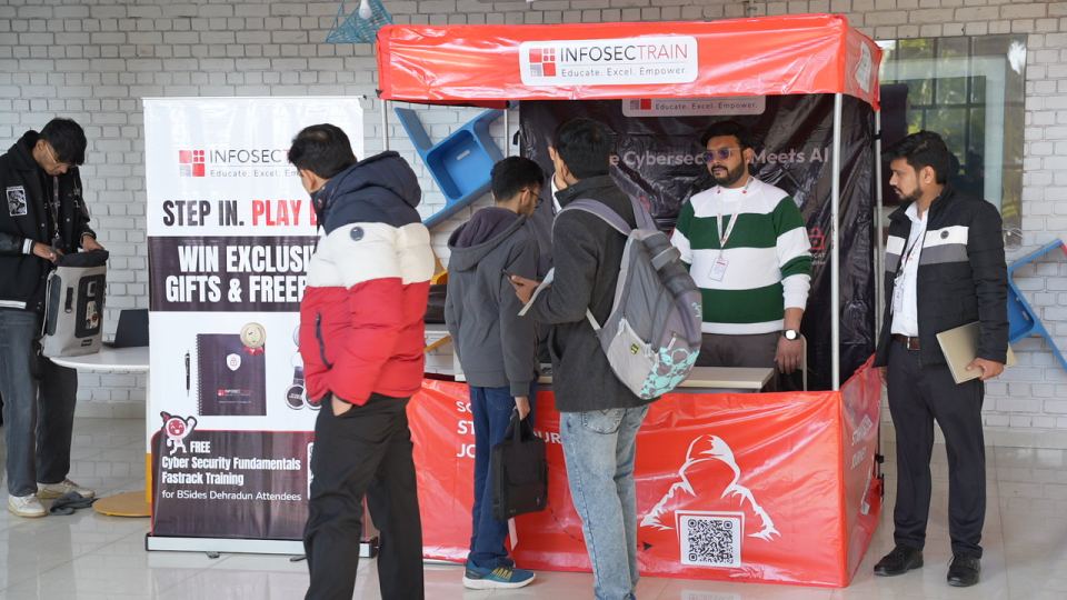 A red pop-up stand with a sponsor's name printed across its canopy, three people leaning on the counter in front of it talking to somebody standing behind, and a tall printed banner beside the stand offering a free course to attendees.