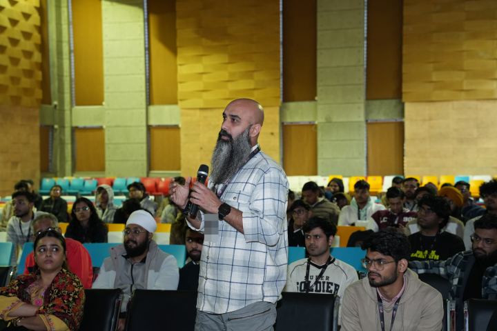 A standing attendee with a long grey beard holding a microphone in both hands, mid-question, in a hall of teal and orange tip-up seats with around twenty-five other people seated around and behind them.