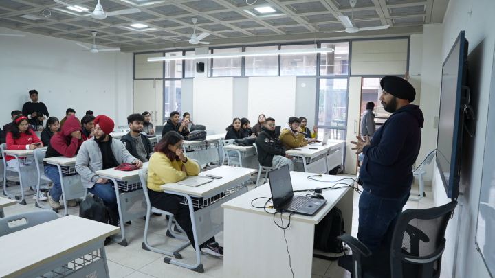 A bright room with daylight along one wall. Someone stands at the front beside a laptop on a desk, mid-sentence with one hand raised, talking to people seated at desks along the window.