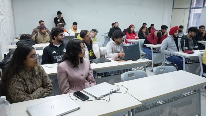 The first desks of a workshop room from the side. Four people sit along a bench with a laptop open between two of them, all four looking up and past the camera towards the front of the room.