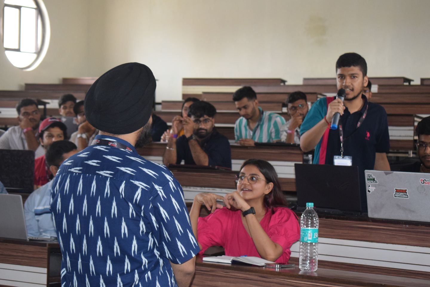 A tiered lecture theatre part-way through a session. Someone in the middle rows is on their feet with a microphone, asking a question. In the front row another person turns and looks up at them, laughing, and the rows behind are full of people at open laptops on long wooden benches.