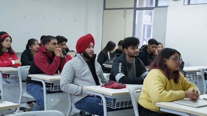 A row of desks photographed along its length, so the people at it are seen one behind another in profile. Laptops and notebooks are open in front of them and all of them are looking left, towards the front of the room.