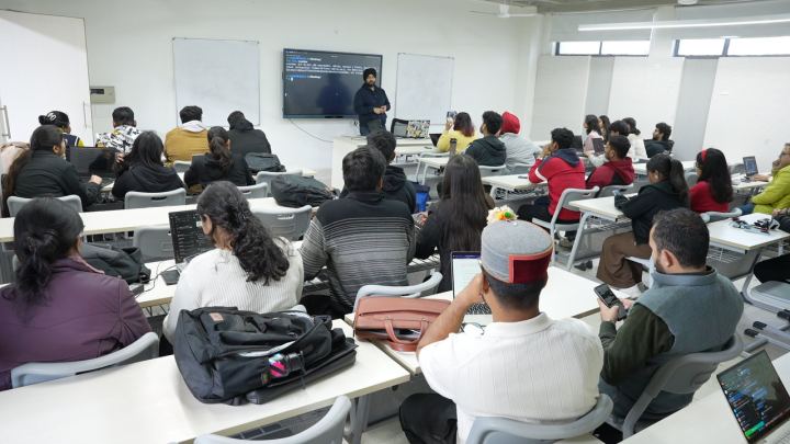 The same kind of room, with a terminal filling the wall screen at the front. A command has just been run and its output is scrolling. People sit at desks in front of it, several with their own laptops open at the same prompt.