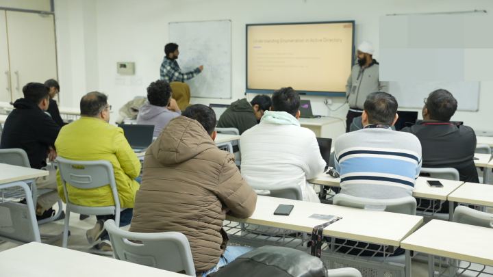 A classroom seen from the back row. At the front, someone stands at a whiteboard covered in a hand-drawn diagram and reaches up to point at it, next to a wall screen showing a slide headed Understanding Enumeration in Active Directory. A dozen people sit at desks between the camera and the board, coats on.